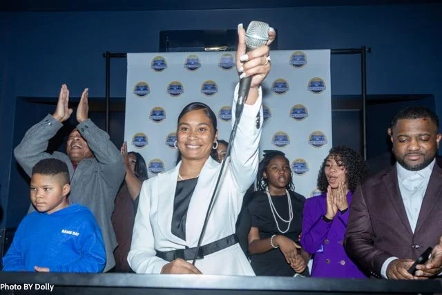 A woman in a white blazer holding a microphone raises her hand, smiling, at an event with other people on stage, some clapping and some taking photos, with a branded backdrop in the background.