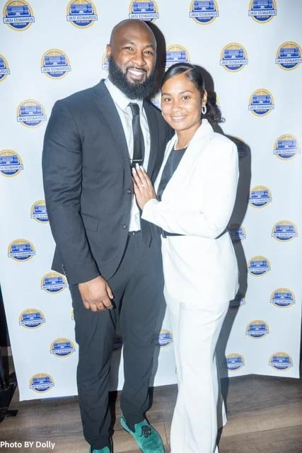 A man and a woman dressed in formal attire standing together in front of a step and repeat backdrop with a logo, smiling at the camera.