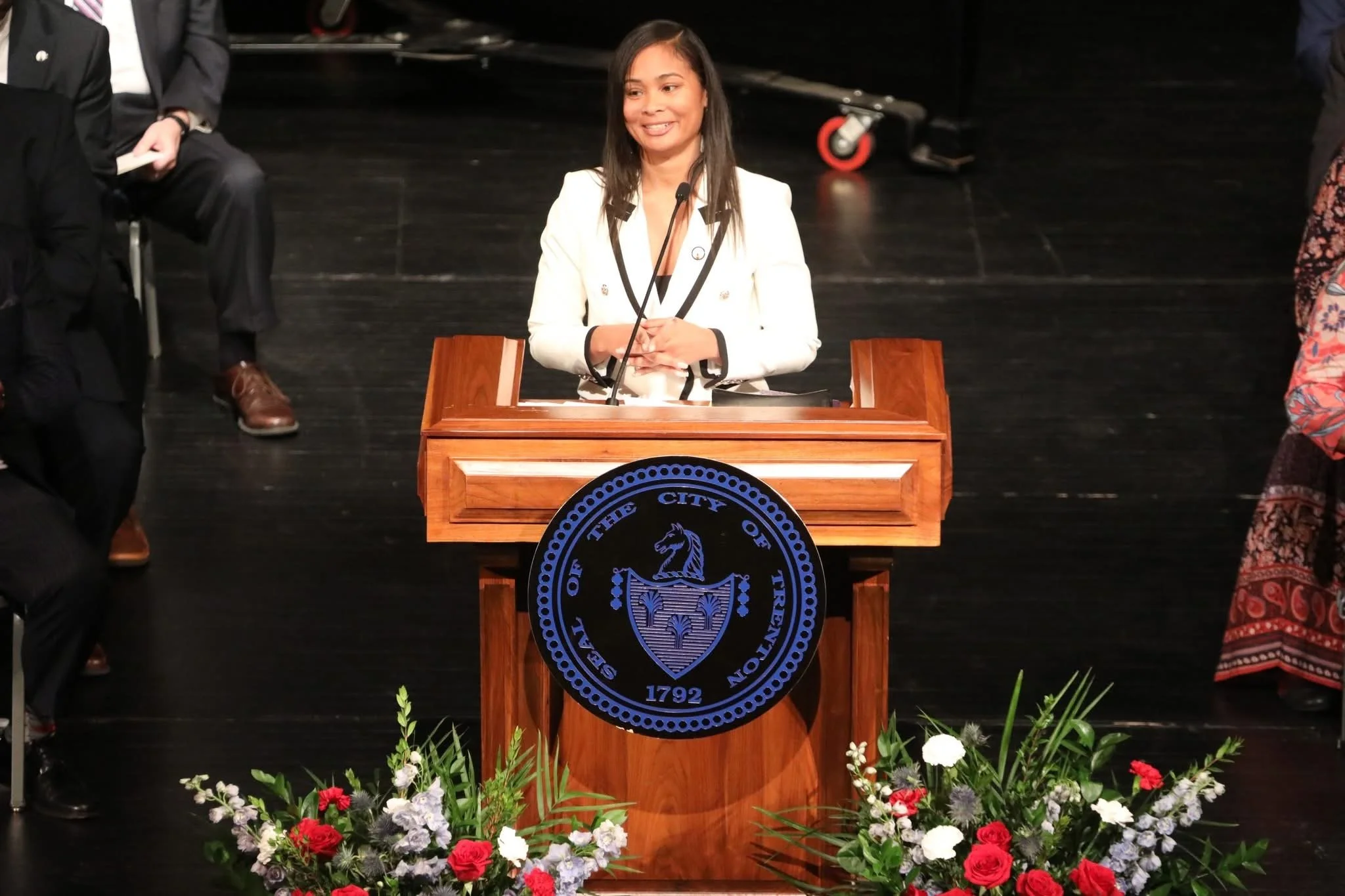 Woman speaking at a wooden podium with the seal of the city of Trenton, surrounded by a floral arrangement, in a formal setting.