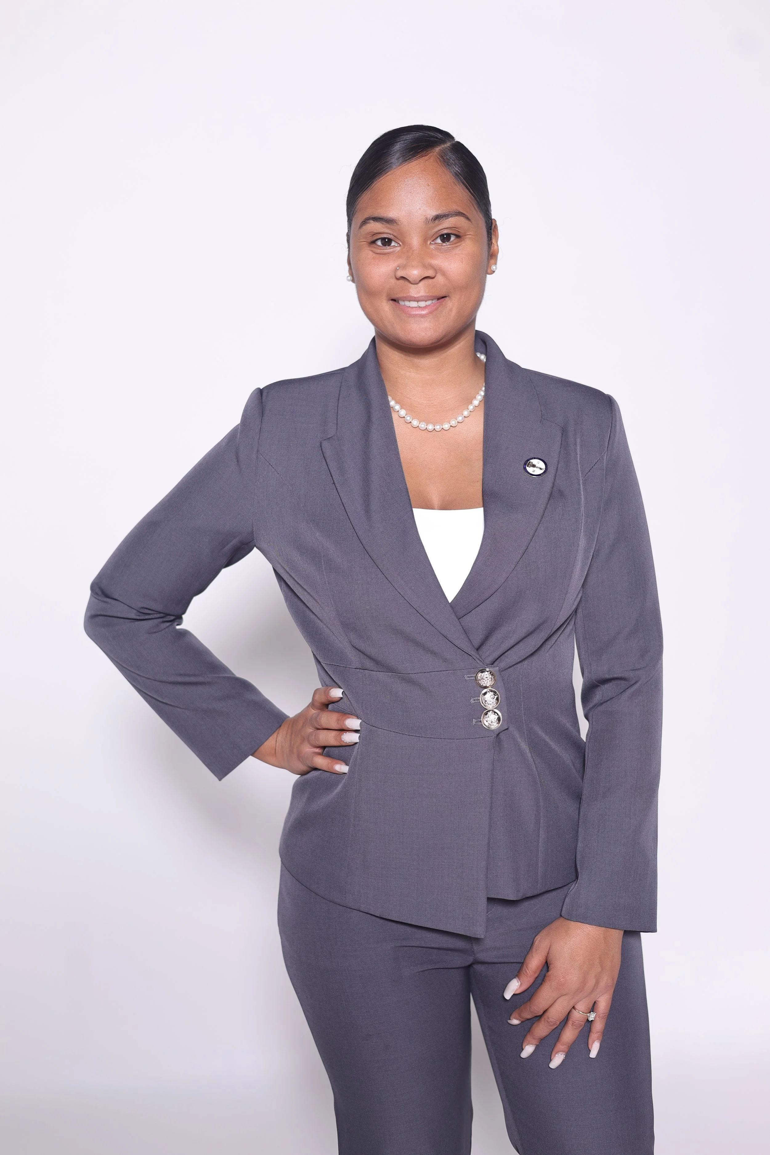 A woman in a gray business suit with a pearl necklace stands against a plain white background, smiling confidently.
