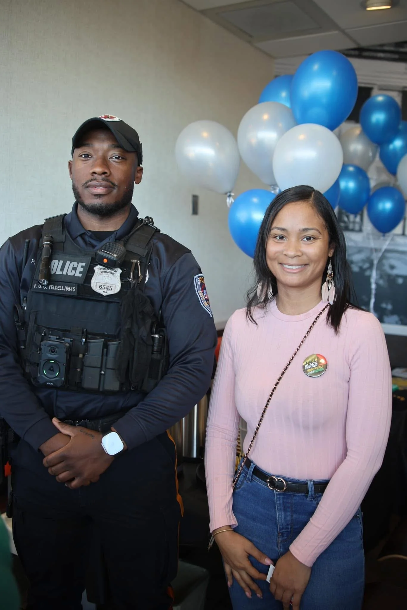 A City of Trenton police officer standing next to Jasi Edwards with balloons in the background. Jasi is smiling and wearing a pink sweater with a 'Black Lives Matter' badge.