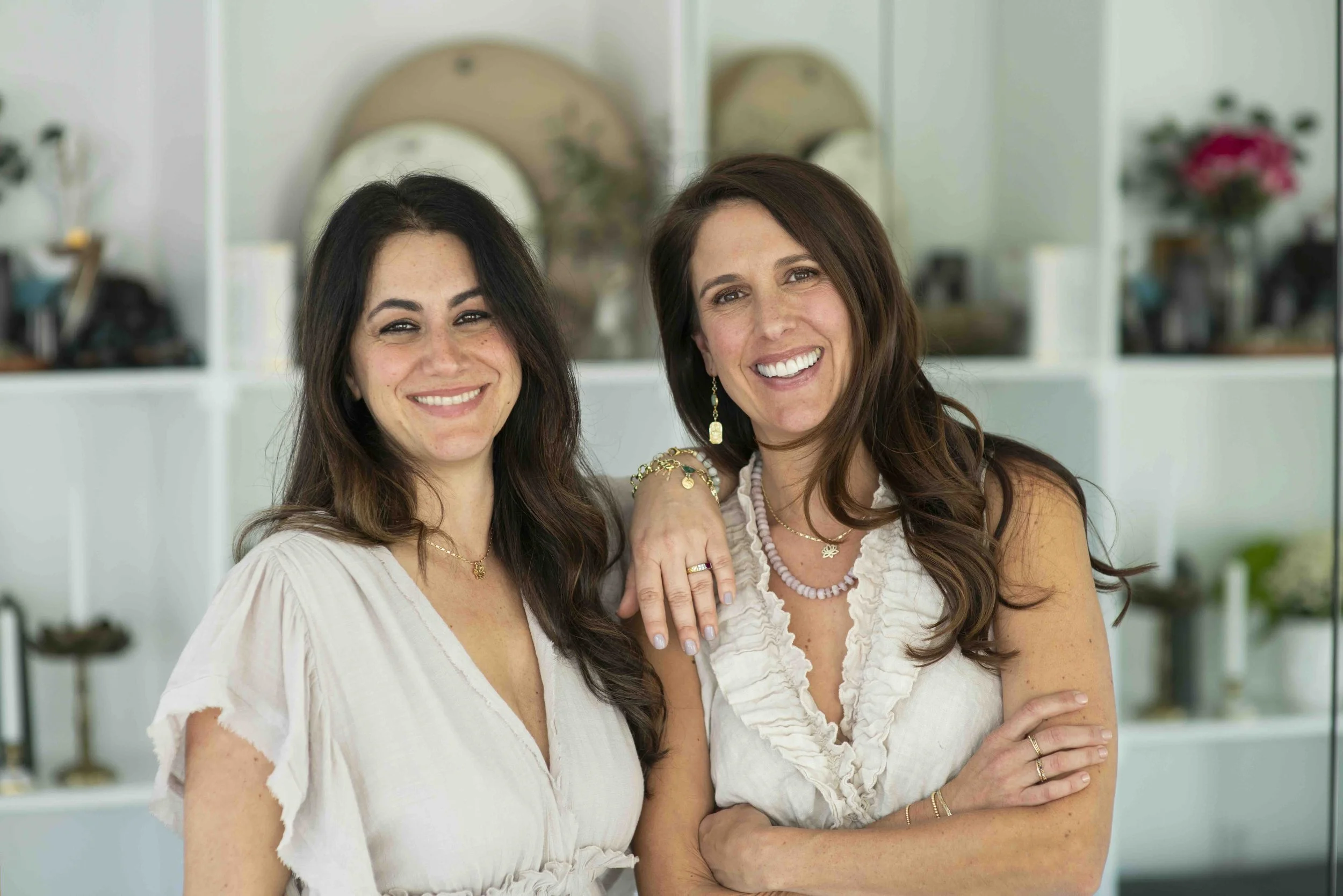 Two women smiling indoors, one with dark hair and the other with light brown hair, wearing light-colored tops, with a white shelf filled with decorative items and flowers in the background.