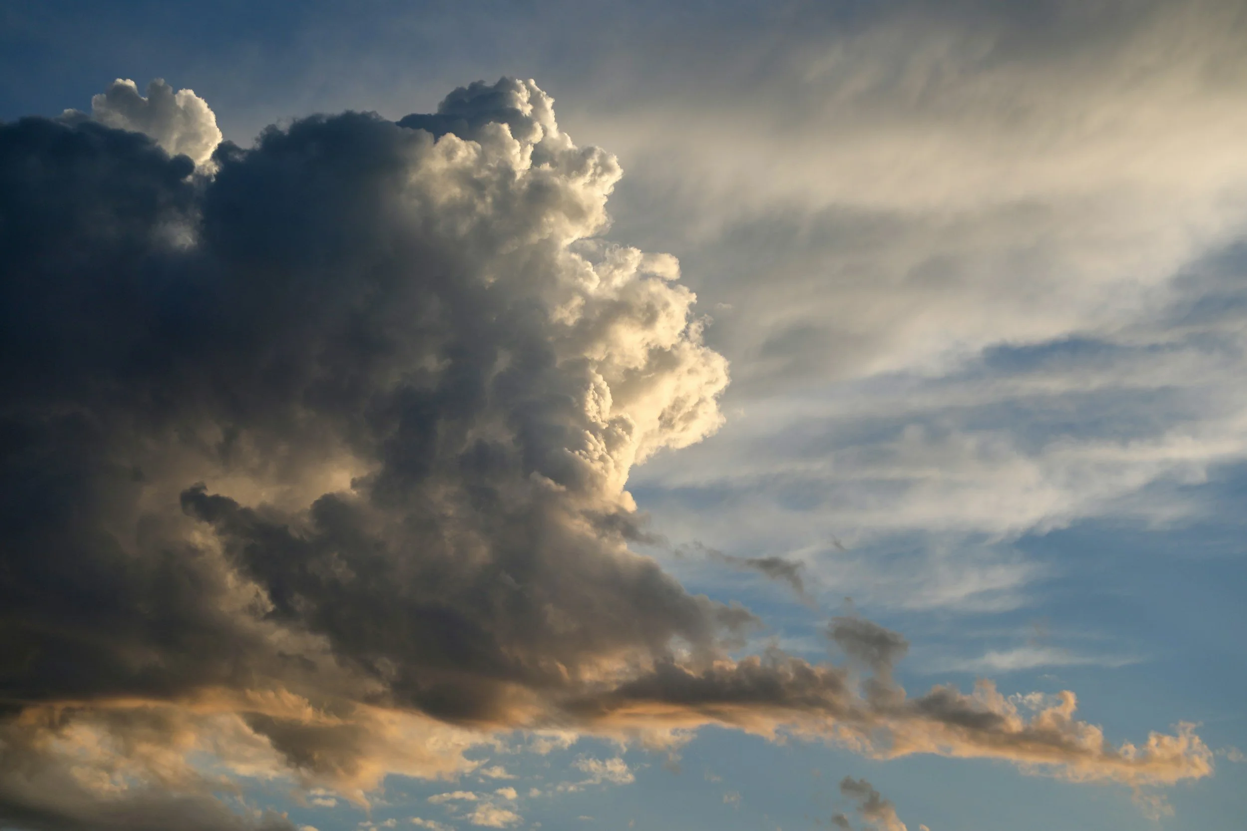 A sky filled with large, dark gray cumulonimbus clouds with some sunlight shining through, and wispy high-altitude cirrus clouds.