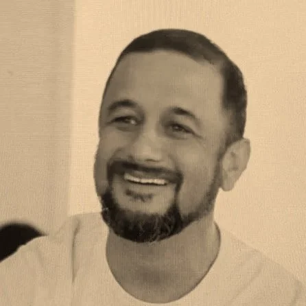 A smiling man with short dark hair and a beard and mustache, wearing a light-colored shirt.