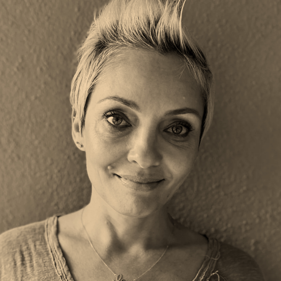 Close-up portrait of a woman with short, spiky blonde hair and light skin, smiling slightly, against a textured wall.