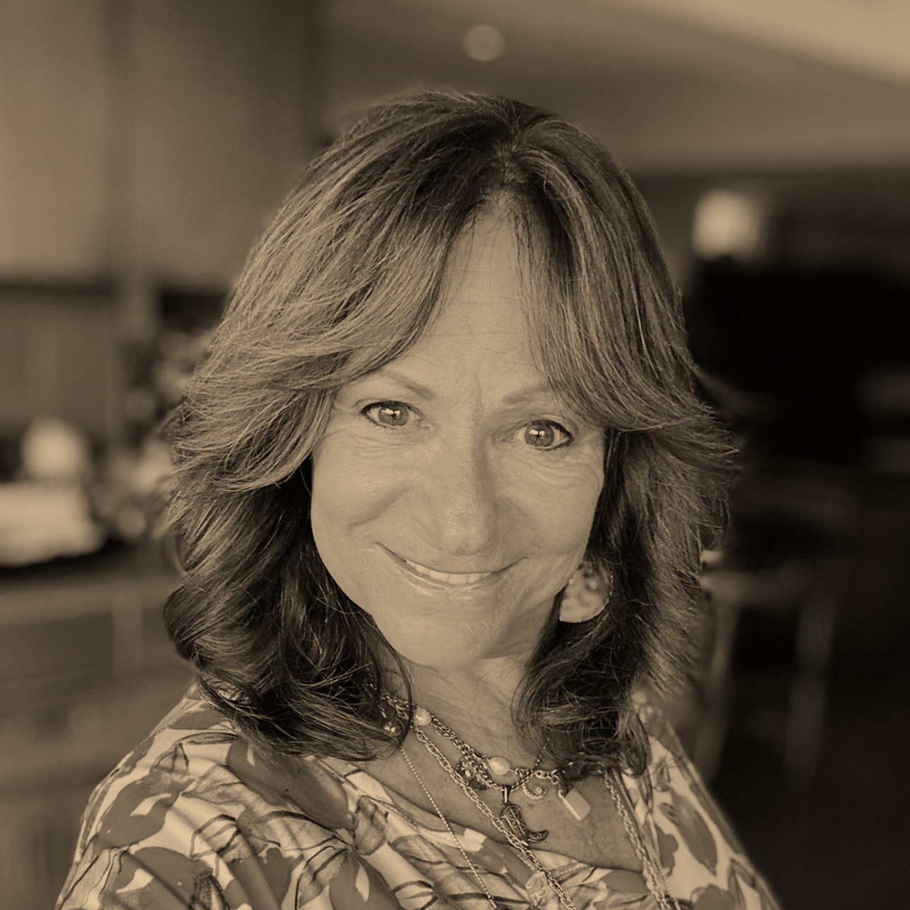A smiling woman with shoulder-length wavy hair and jewelry, in a room with a blurred background.