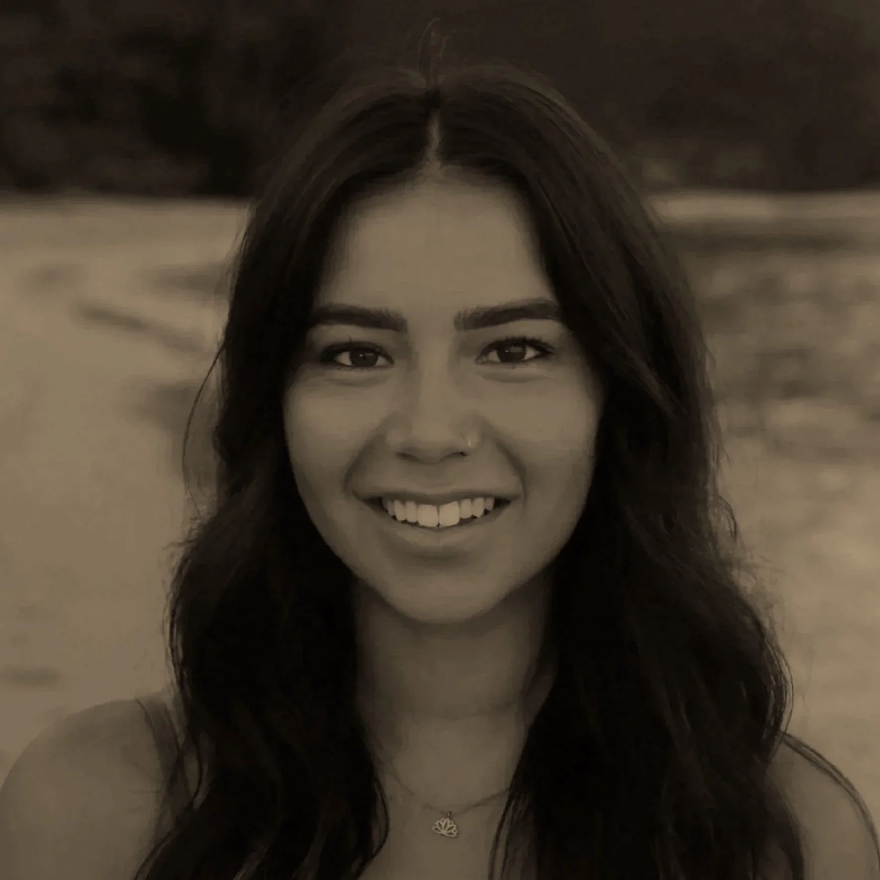 A woman with long dark hair smiling outdoors, with a blurred background of water and trees.