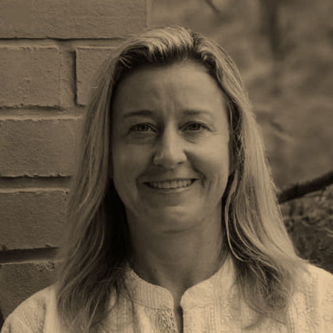 A woman with long blonde hair smiling at the camera, standing next to a brick wall.
