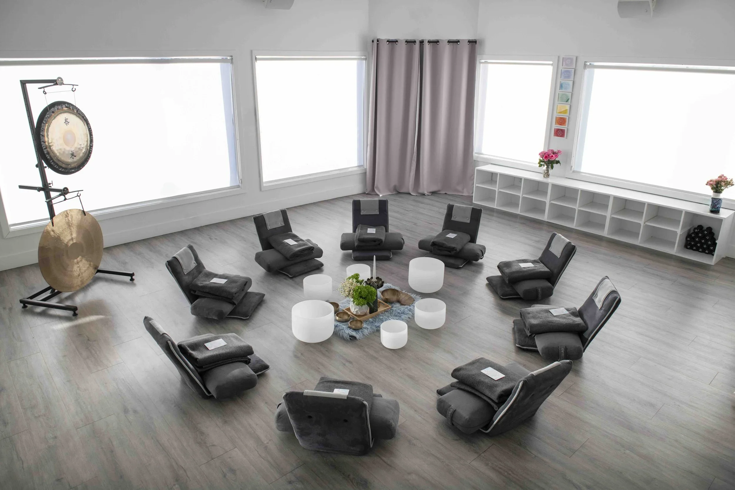 A circular arrangement of nine gray chairs with black cushions in a bright, spacious room with large windows, a gong and cymbals on the left, a white shelf with pink flowers and colorful art on the right, and a central flower arrangement with candles and decorative objects on a small table.