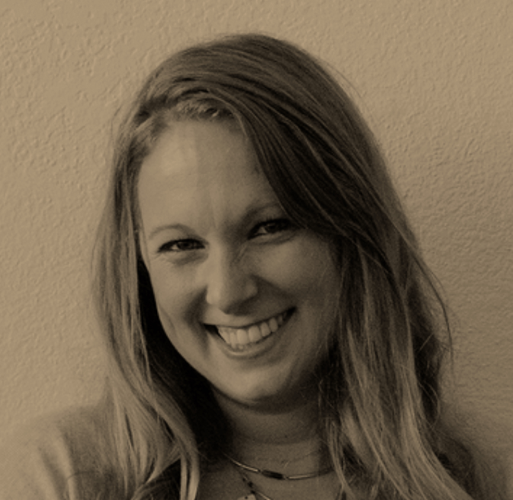 A woman with long, wavy hair smiling at the camera, showing her teeth, with a light-colored wall in the background.