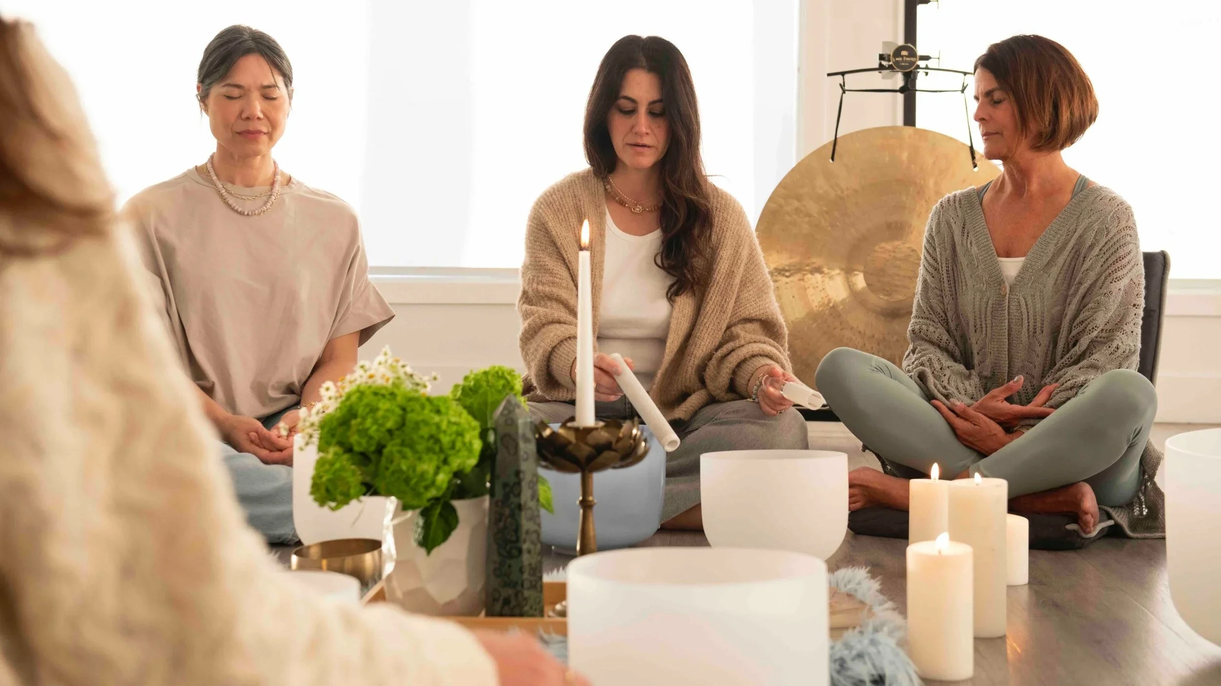 Three women sitting cross-legged and meditating in a peaceful, softly lit room with candles, flowers, and a gong in the background.