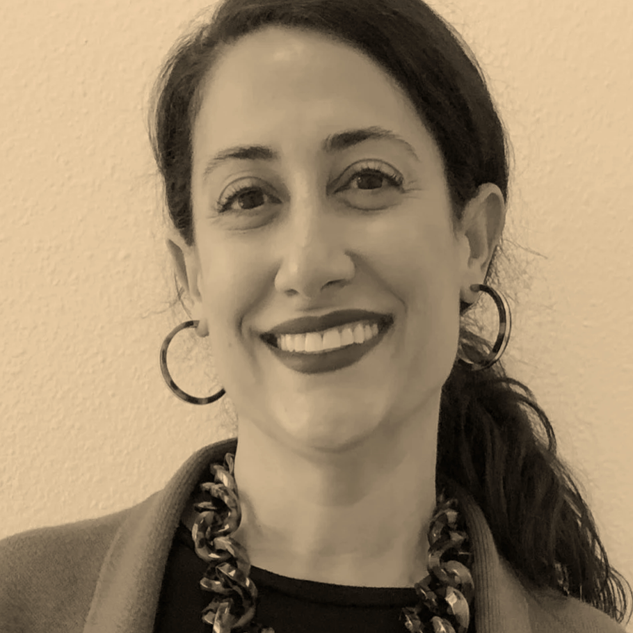 A woman smiling with dark hair, wearing hoop earrings and a chunky necklace, standing against a plain wall.