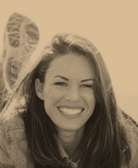 A woman with long hair smiling and showing her teeth, with wind blowing her hair, and a blurred background featuring a partially visible bicycle wheel.