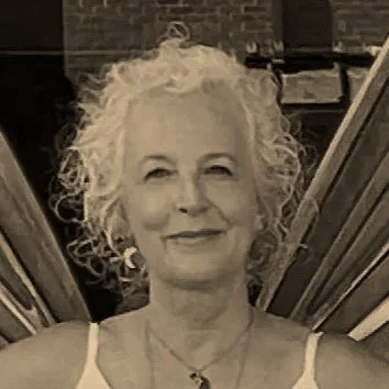 A woman with curly white hair smiling at the camera, standing in front of a staircase.