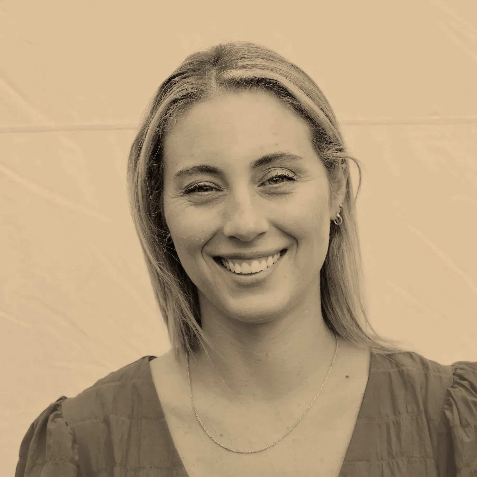 A smiling woman with blonde hair, wearing earrings, a necklace, and a dark top, standing against a plain background.