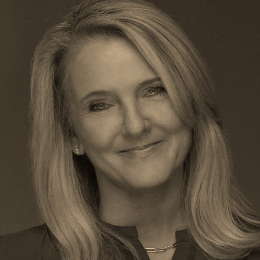 A woman with long blonde hair and a gentle smile, wearing a dark top and a chain necklace, posing against a dark background.