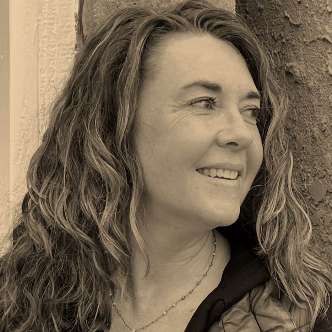 A woman with long wavy hair smiling and looking to the right, standing outdoors near a textured wall.