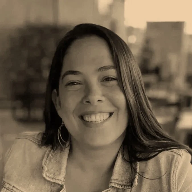 A smiling woman with shoulder-length dark hair, wearing hoop earrings and a denim jacket, in a warmly lit indoor setting.