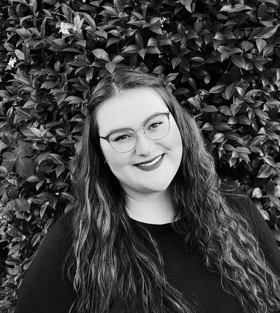 A woman with long wavy hair and glasses smiling in front of a leafy background.
