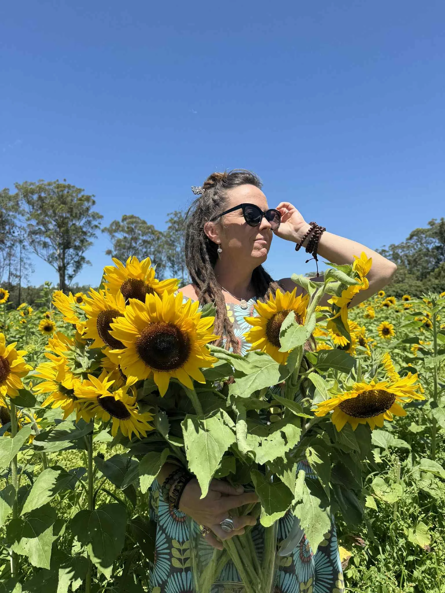 Woman with sunglasses and dreadlocks standing in a sunflower field, holding a large bouquet of sunflowers, under a clear blue sky.