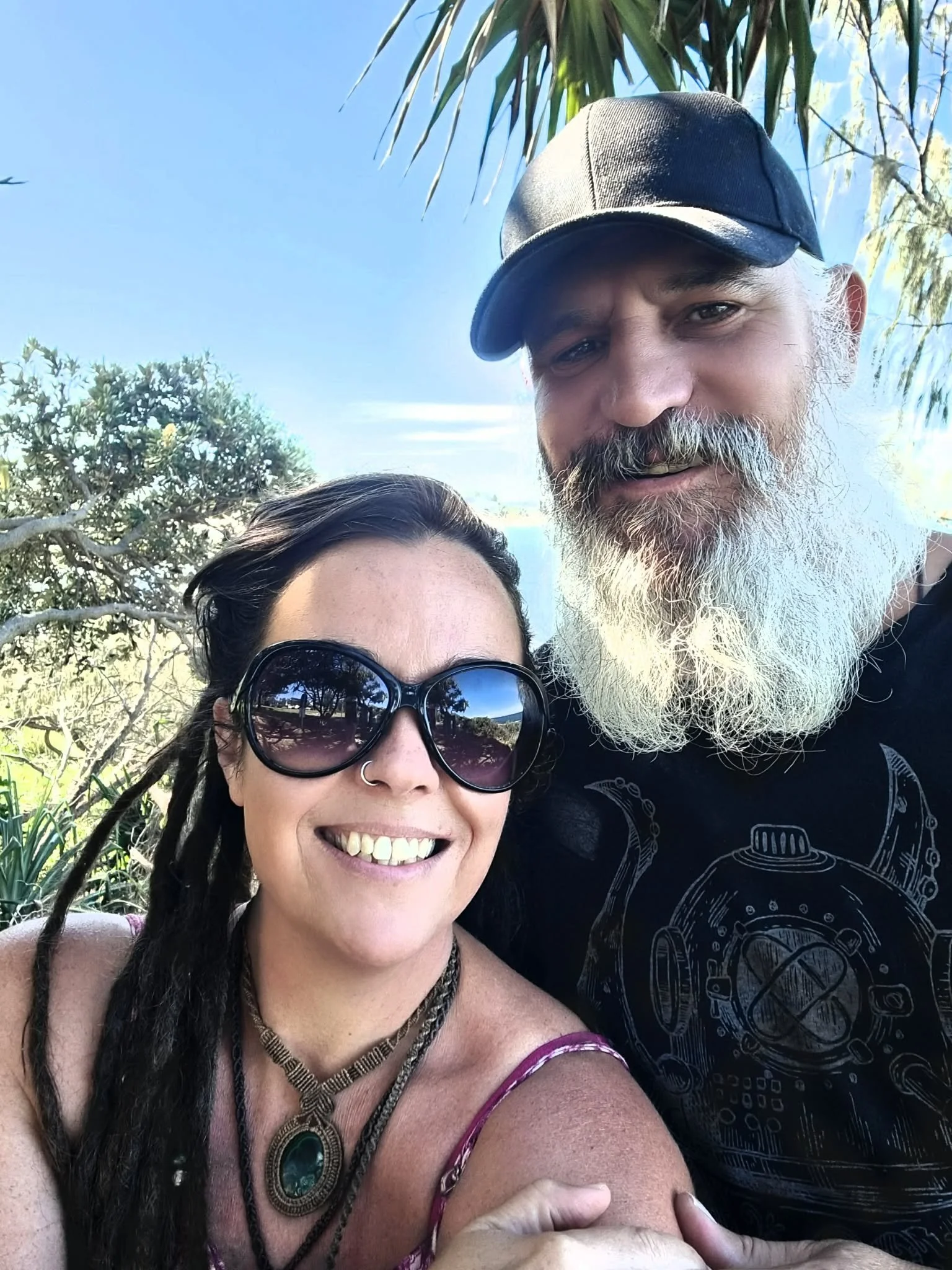 A woman with sunglasses and a necklace smiling, taking a selfie with a man with a gray beard and a black cap outdoors under blue sky and tropical trees.