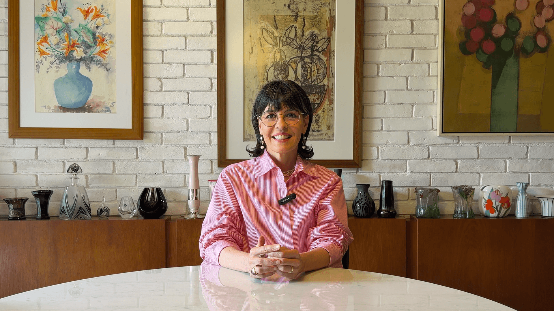 Mulher sorridente vestido camisa social rosa clara, com mãos cruzada sob uma mesa branca redonda e com microfone de lapela preto à direita da camisa. Ao fundo, três quadros de arte, acima de uma estante com vasos de vidro coloridos.