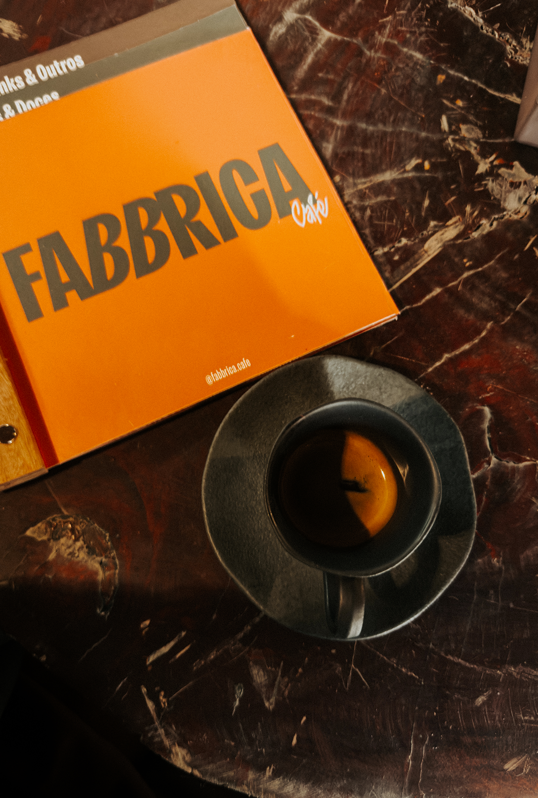 Papel laranja com escrito Fabbrica Café em verde escuro, sob mesa de mármore rajado preto e laranja, acima de uma xícara verde com café preto dentro, repousando sob um pires verde.