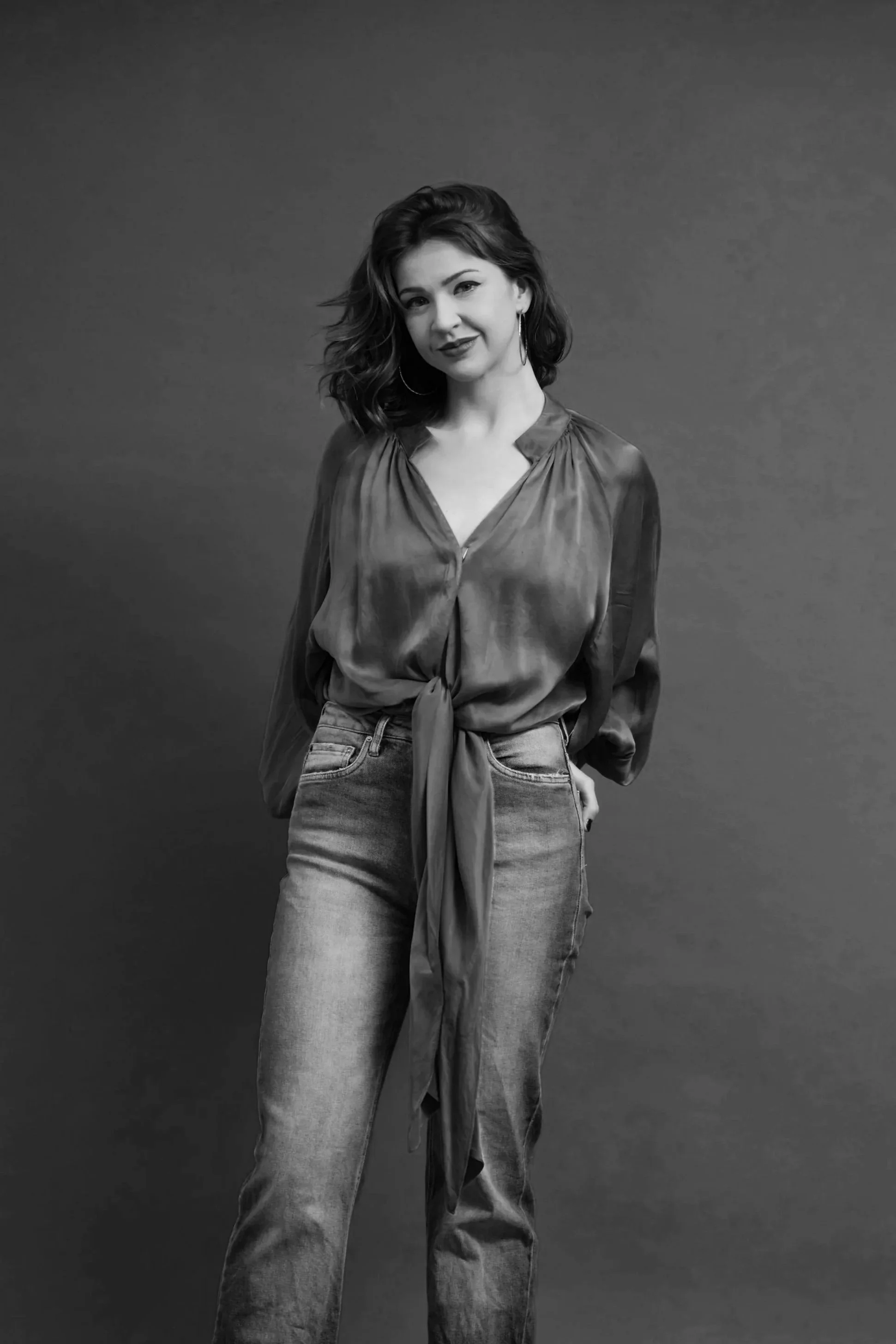 Black and white portrait of a woman with wavy hair, wearing a satin blouse tied at the waist and jeans, standing against a plain background.