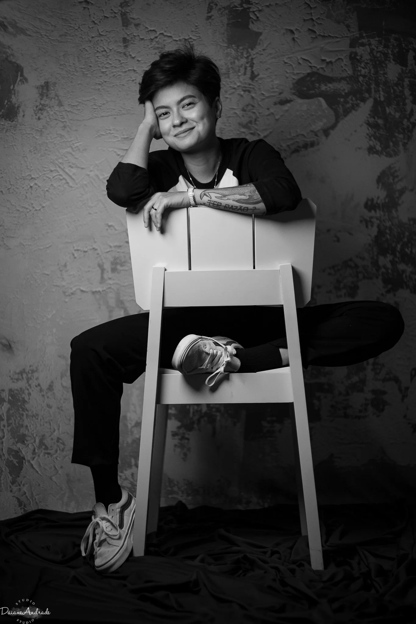 A young woman with short hair, smiling, sitting on a tall chair with one leg crossed over the other, resting her arm on the back of the chair. She is wearing sneakers, dark pants, and a long-sleeved shirt. The background is textured, and the photo is in black and white.