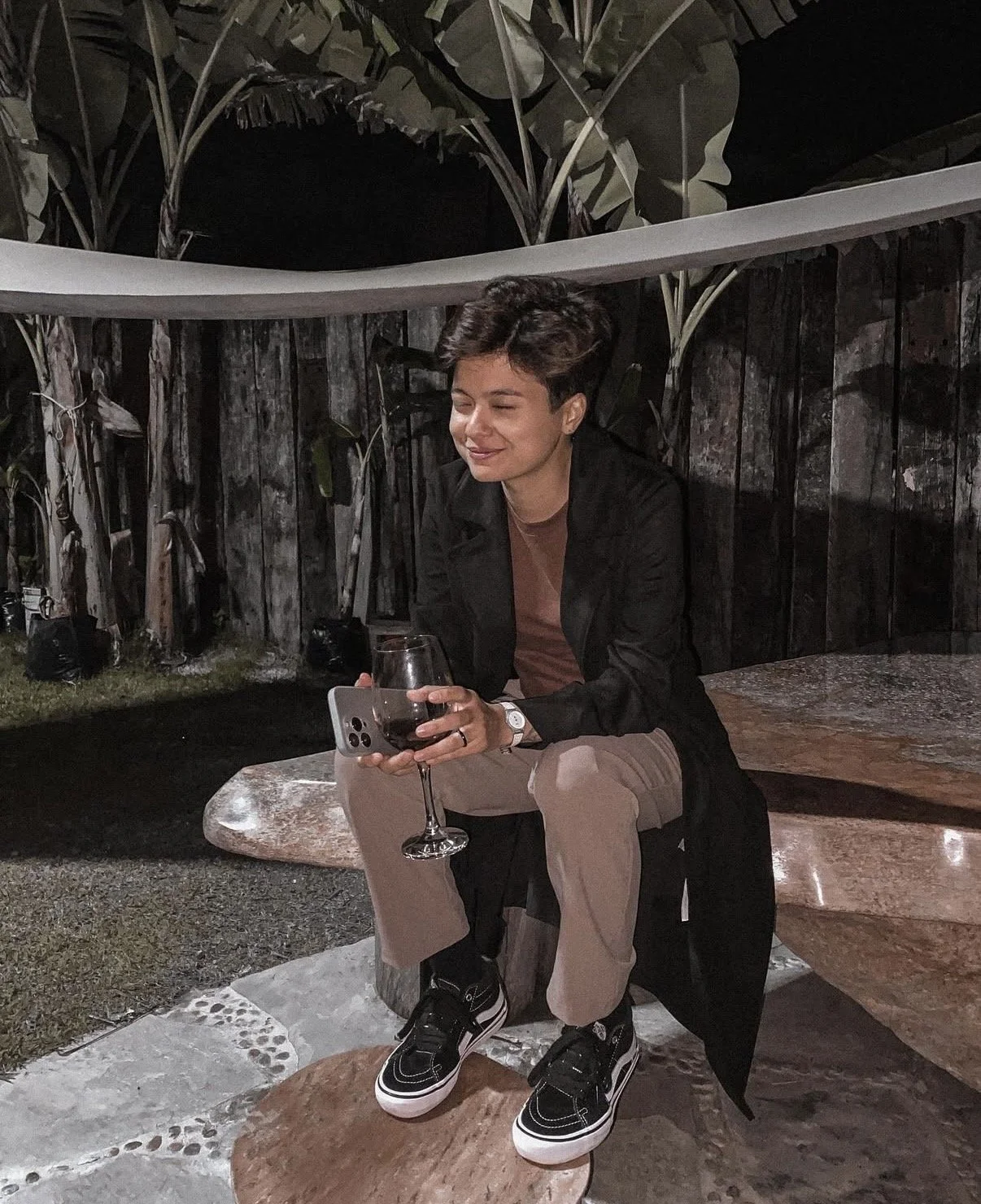 Woman sitting outdoors at night, holding a glass of red wine, wearing a black jacket, brown pants, and black sneakers, with a brick and wooden fence in the background.