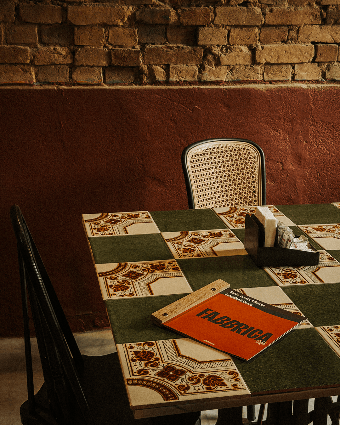 A table with a colorful tiled surface in a restaurant, with a black container holding napkins, sugar packets, and utensils, and a menu titled "FABRIC" on the table. There are chairs around the table, one with a woven cane backrest and another partially visible, against a brick and red painted wall.