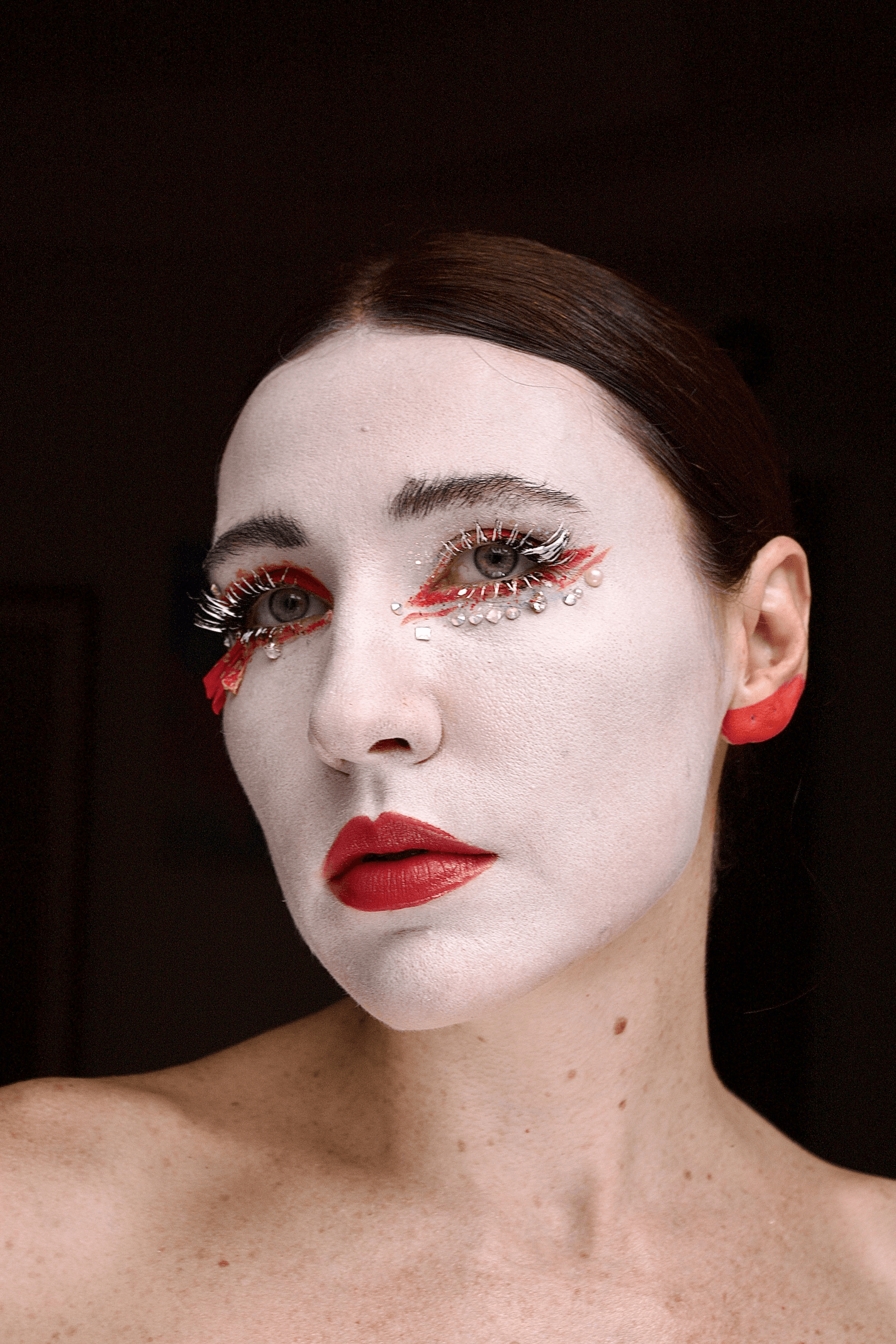 Mulher em close-up com maquiagem artística. Rosto coberto por base branca, lábios vermelhos marcantes, delineado vermelho gráfico ao redor dos olhos, cílios brancos dramáticos e aplicações de pedrarias abaixo dos olhos
