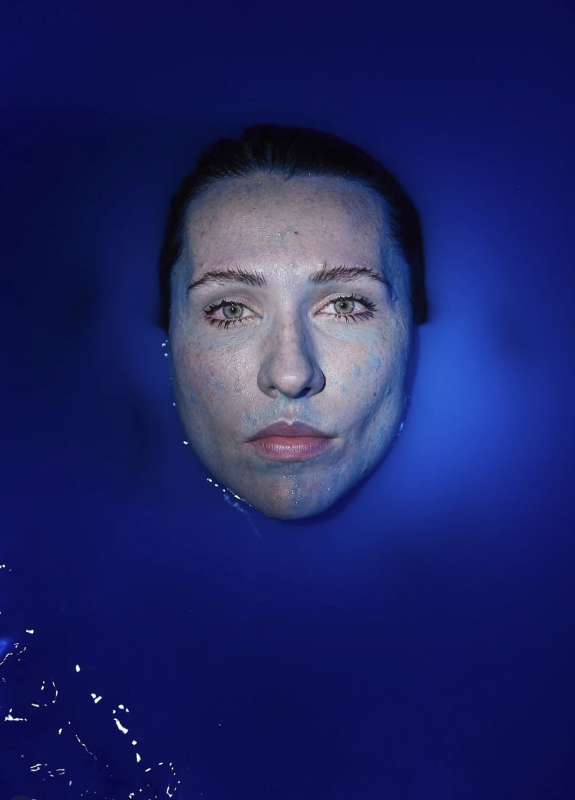 Rosto de mulher emergido de um líquido azul intenso, com expressão neutra e olhar direto para a câmera.