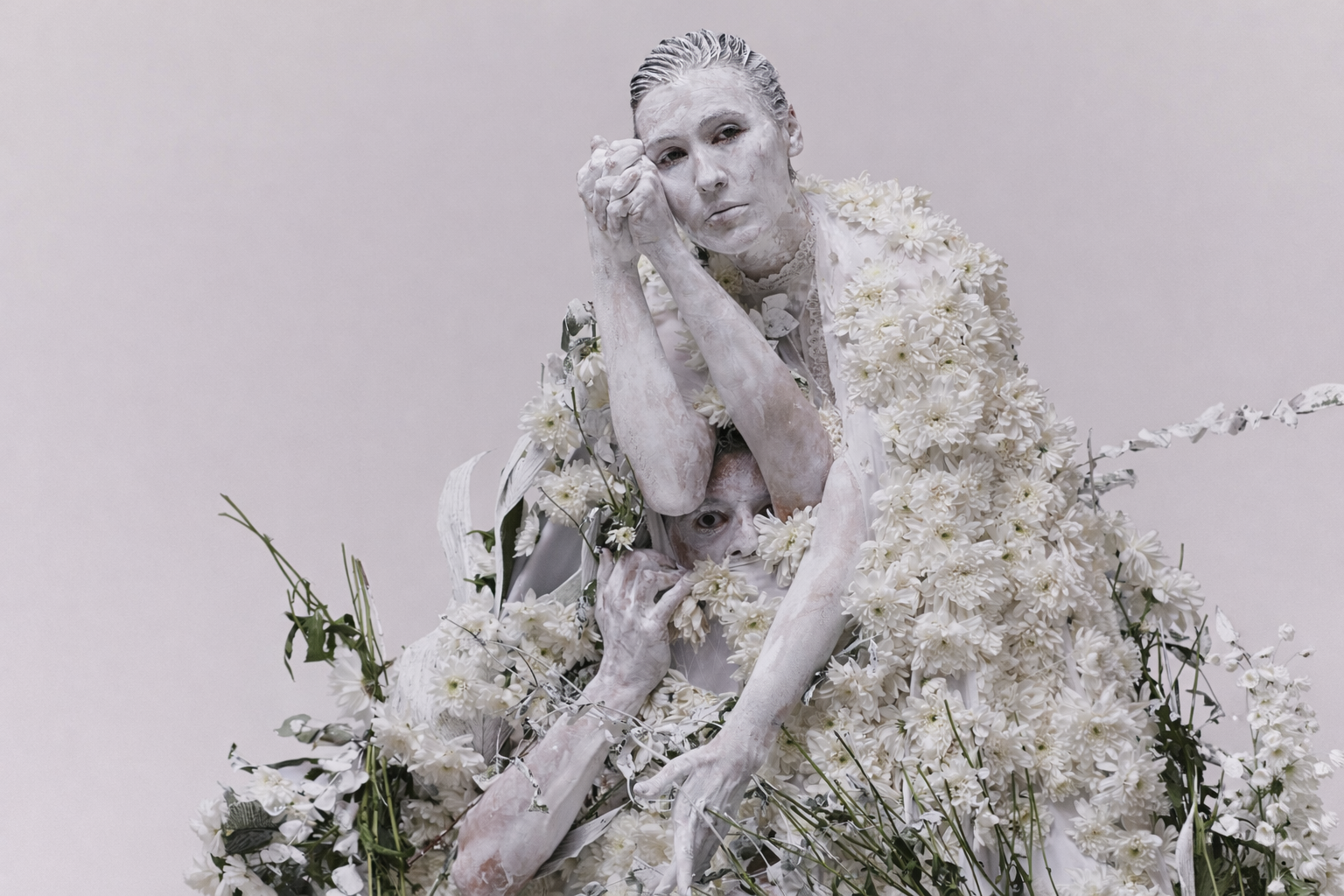 Two people painted white, surrounded by white flowers; one rests their head on their clasped hands, while the other peeks through the flowers, creating an artistic and surreal scene.