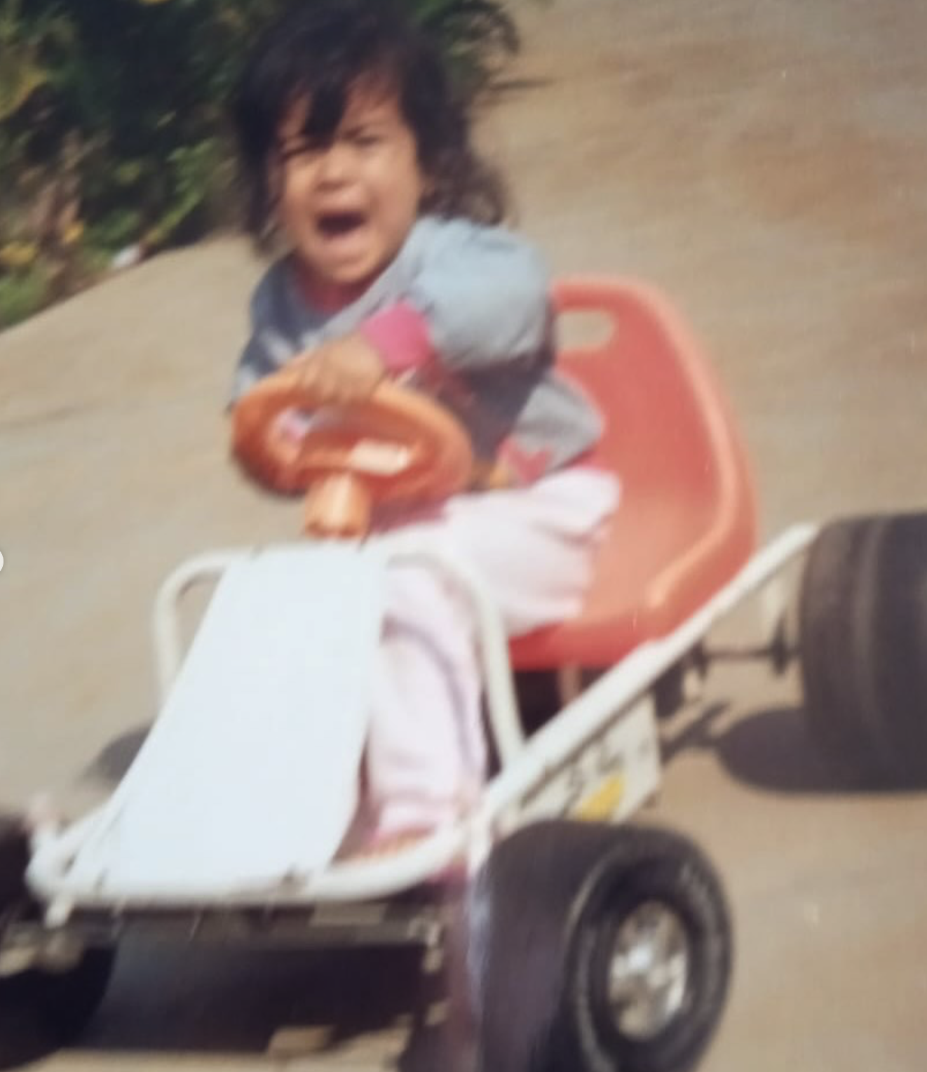 Child with dark hair in a gray shirt, sitting in a small go-kart with a red seat, holding the steering wheel with a big smile and open mouth.