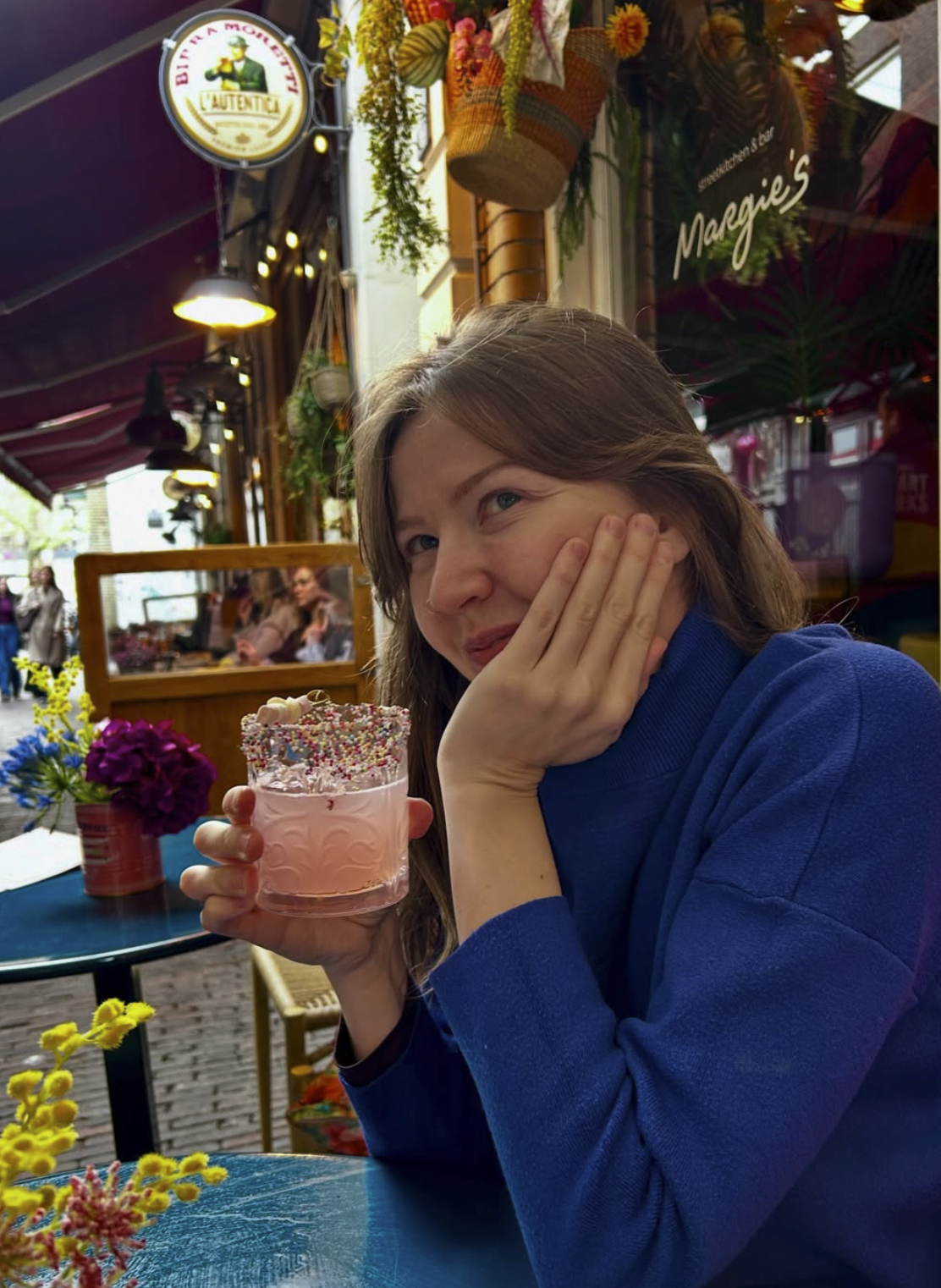 A woman in a blue sweater sitting at a colorful outdoor café, holding a pink milkshake with sprinkles, smiling, surrounded by flowers and string lights, with a street scene in the background.