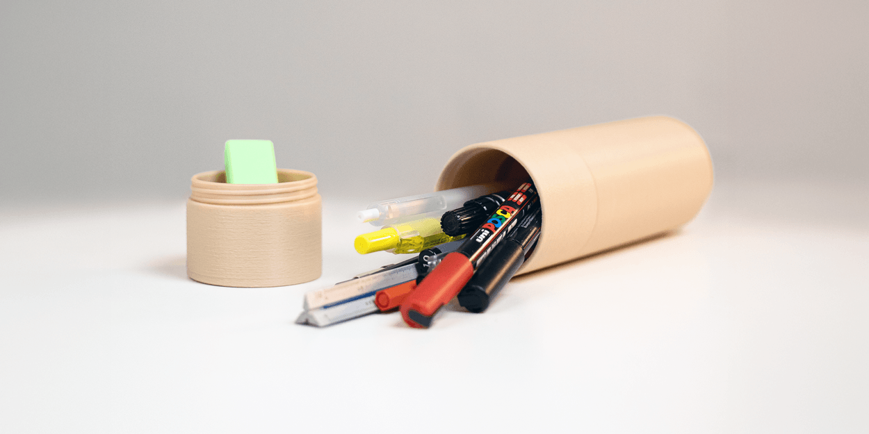 Estojo cilíndrico bege deitado em superfície branca, com canetas, lápis e uma régua parcialmente para fora. Uma tampa bege com uma borracha dentro fica à esquerda do estojo.