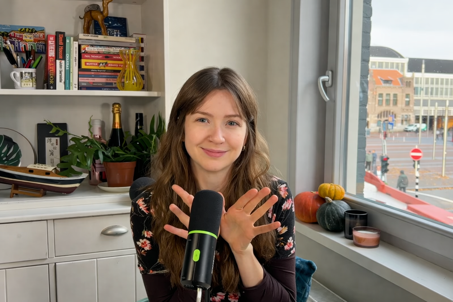 Mulher sorrindo sem mostrar os dentes, usando blusa de manga comprida preta estampada com flores cor de rosa. Mãos abertas, com as palmas frente viradas para a câmera, e os polegares cruzados, atrás de um microfone preto com detalhes verdes.
