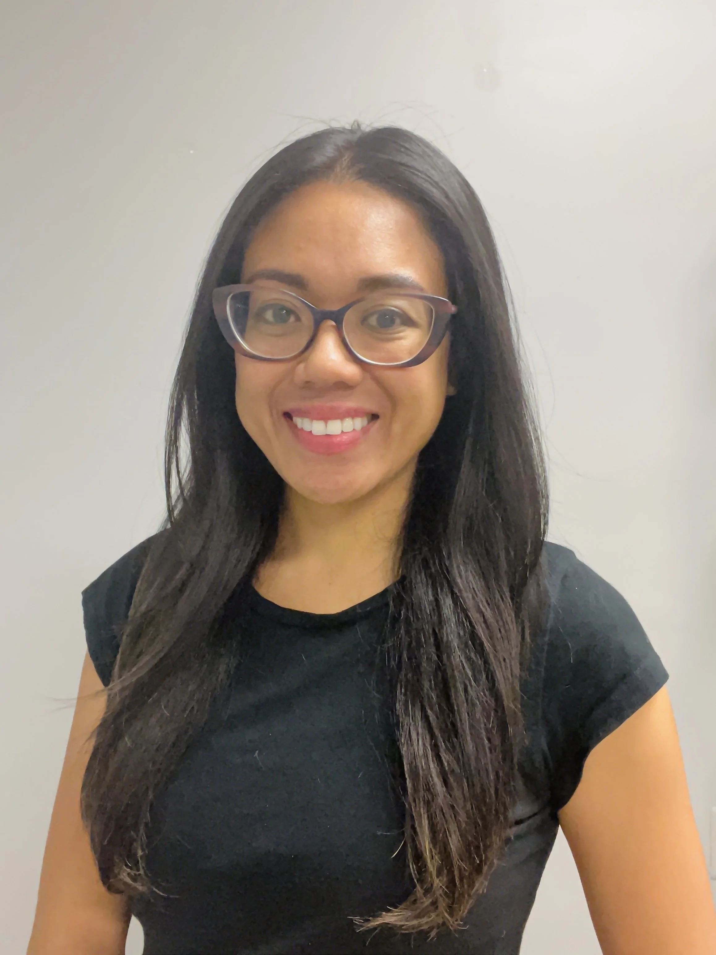 A woman with long black hair, glasses, and a black shirt smiling, standing against a plain light-colored background.