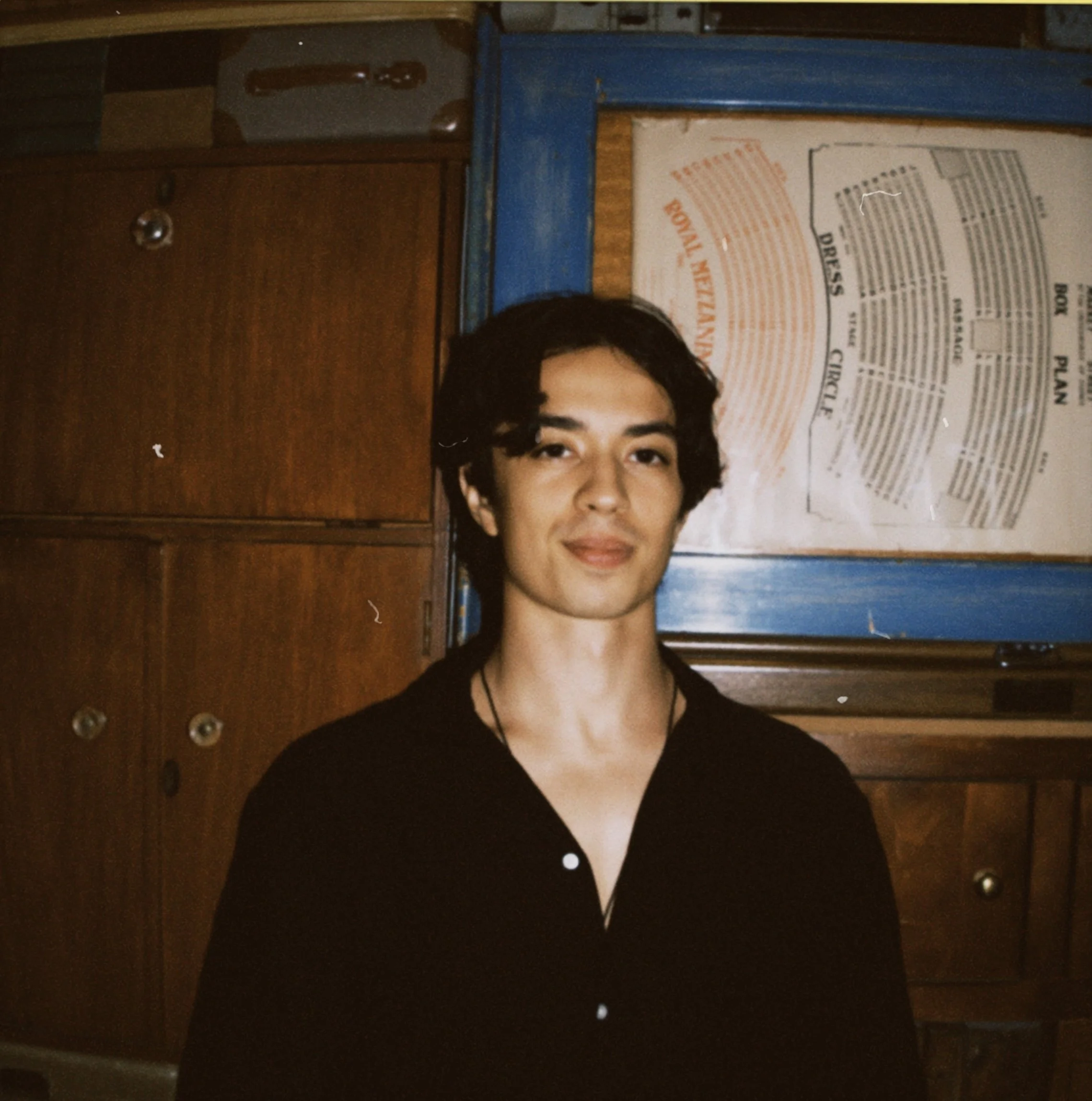A young person with dark, wavy hair and light skin, smiling softly, is wearing a black shirt and is standing in front of a wooden and blue background with restaurant or cafeteria menu signs.