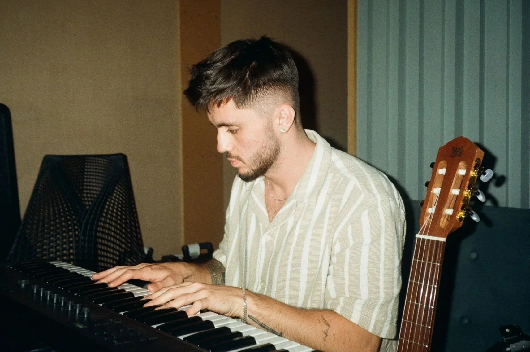 Man with short dark hair and beard playing the piano in a music studio, with an acoustic guitar resting on the chair beside him.