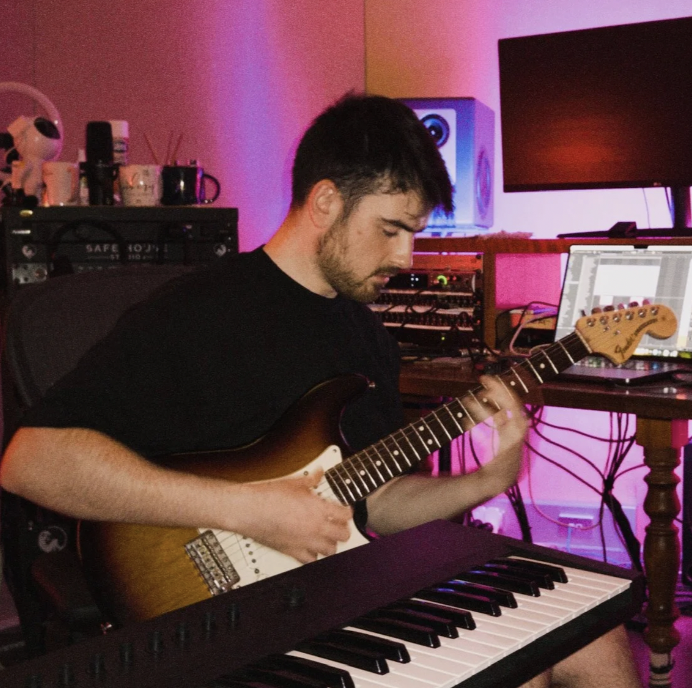 A young man is playing an electric guitar while sitting at a keyboard in a music studio with pink and purple lighting.