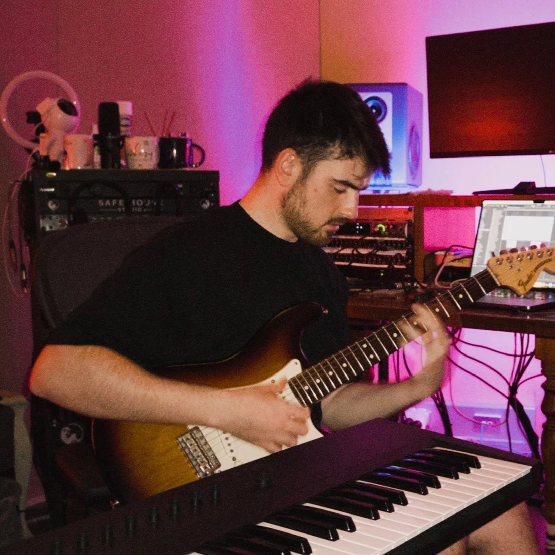 A man smiling and playing an electric guitar in a room with pink and purple lighting, with computer monitors and audio equipment in the background.