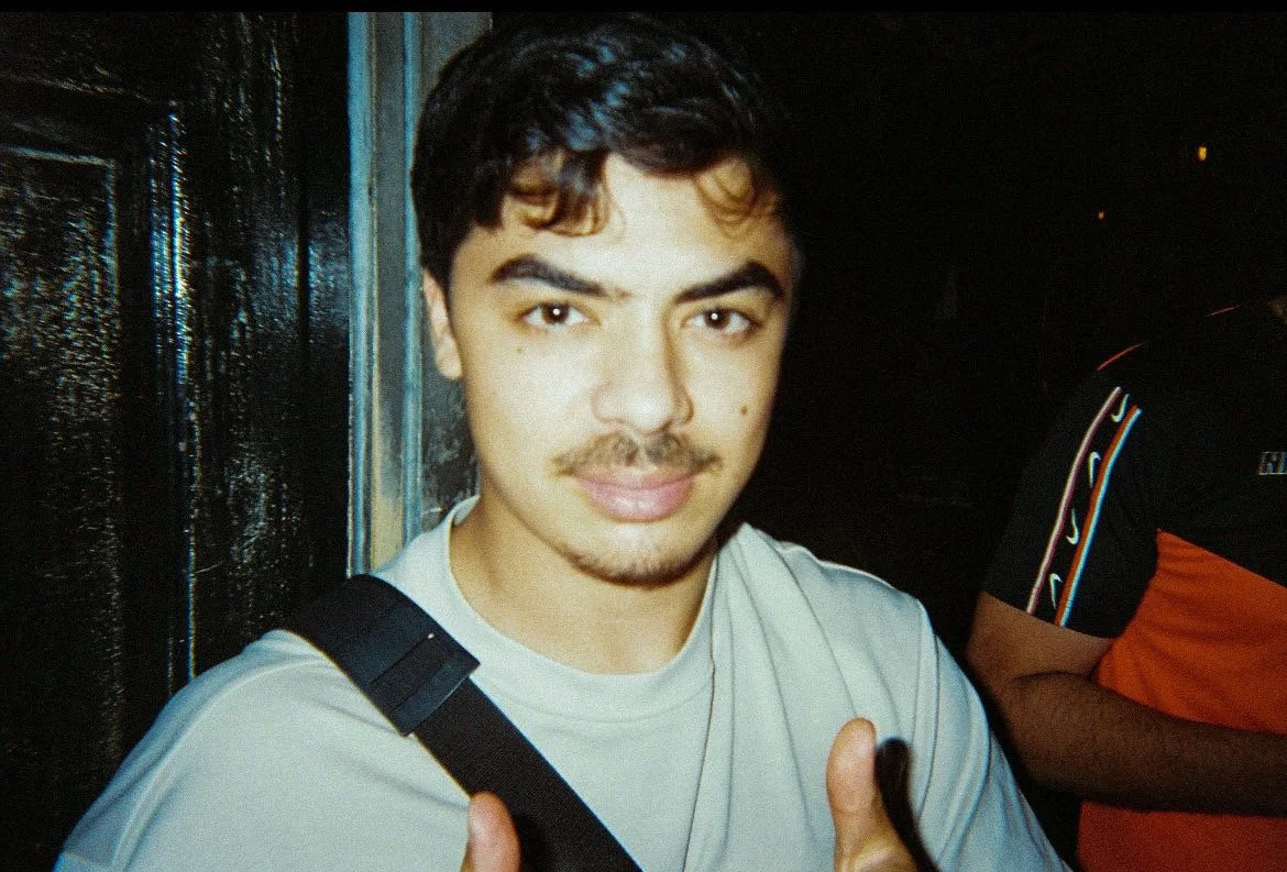 A young man with dark hair, wearing a white t-shirt, giving a thumbs-up. He has a slight mustache and is standing in front of a dark, shiny surface.