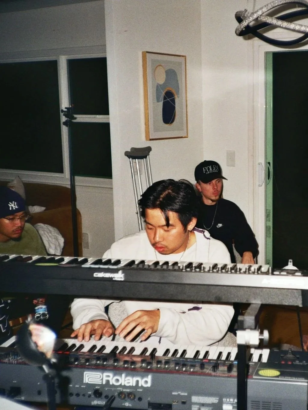 Three young men, two sitting and one standing behind a keyboard, in a casual indoor setting. One is playing a Roland keyboard, a second sitting to the left wearing a Yankees cap, and a third in the background wearing a black cap with 'FOLEY' written on it. The room has white walls, a painting, large windows, and a modern chandelier.