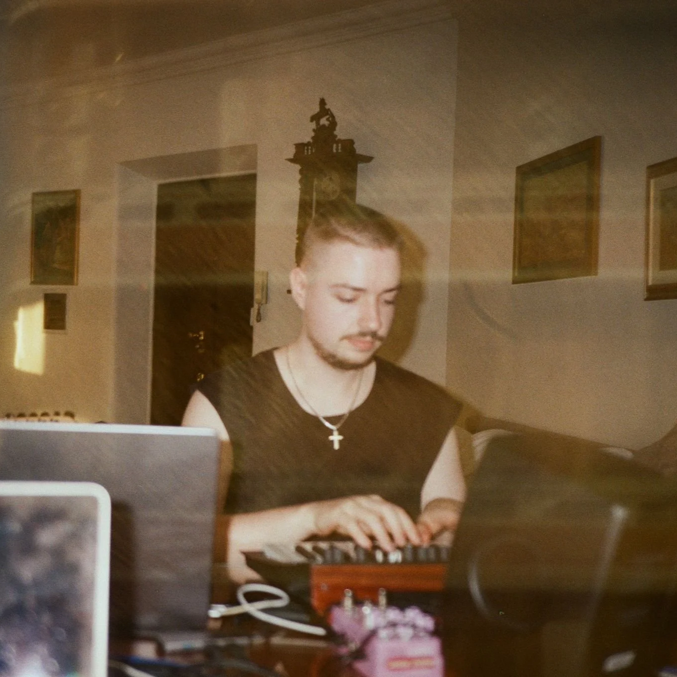 A young man with a goatee and short hair, wearing a sleeveless black shirt and a cross necklace, is working with electronic music equipment on a wooden table in a room with framed paintings and a clock on the wall.