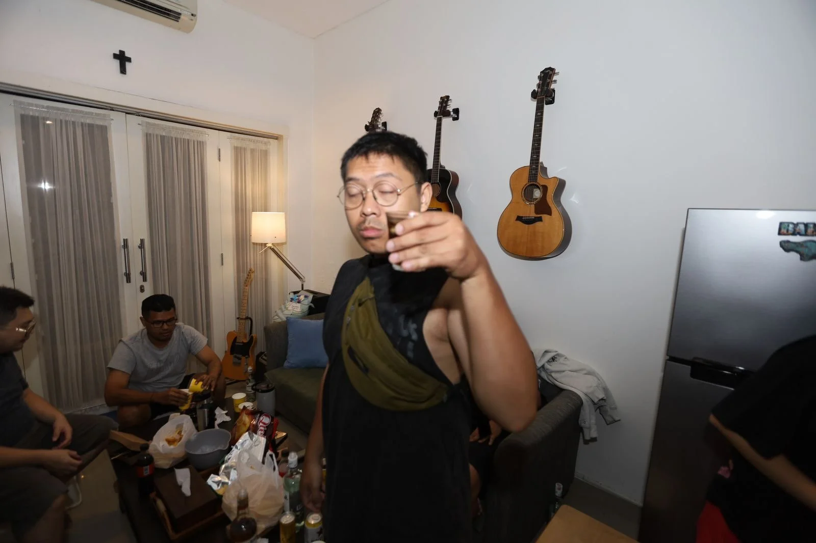 Young man with glasses and short hair raising an empty shot glass, surrounded by friends in a living room with guitars on the wall, a sofa, and a table with food and drinks.