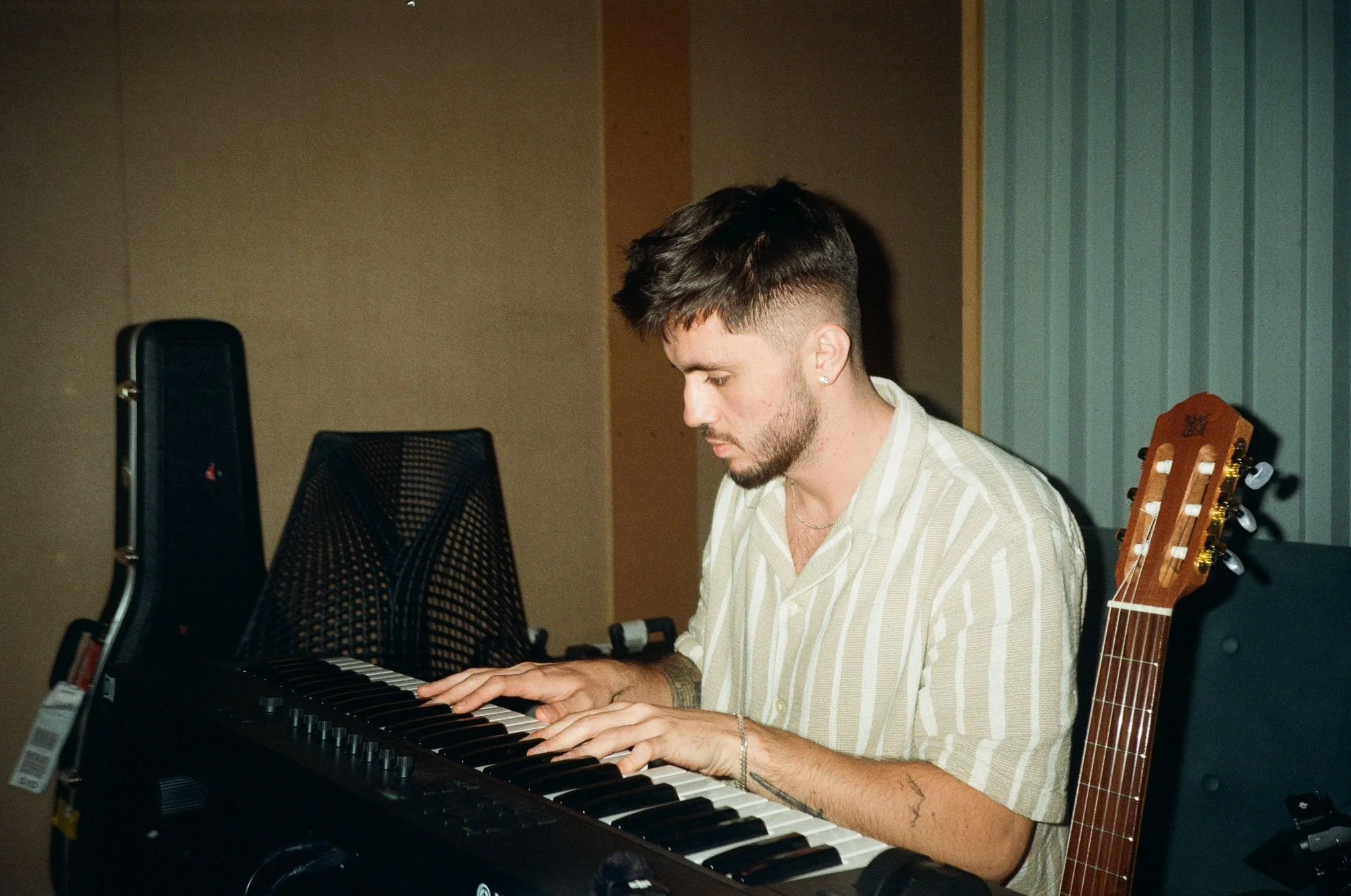 A young man with short dark hair and a beard playing a keyboard in a music studio, with a guitar leaning against the chair next to him.