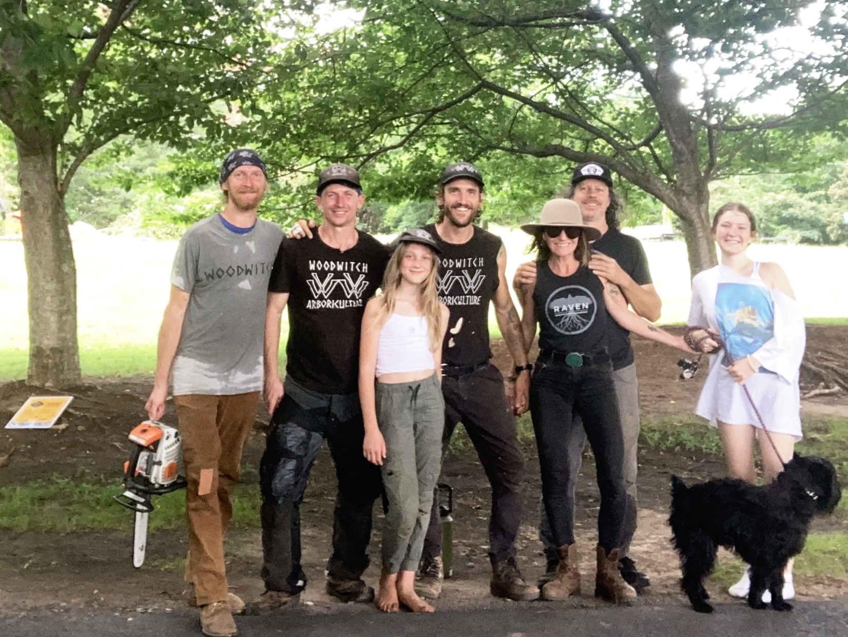 Group of six people and a dog outdoors under a large tree, with some equipment nearby. Woodwitch Arborists, Tree Preservation & Removal in WNC