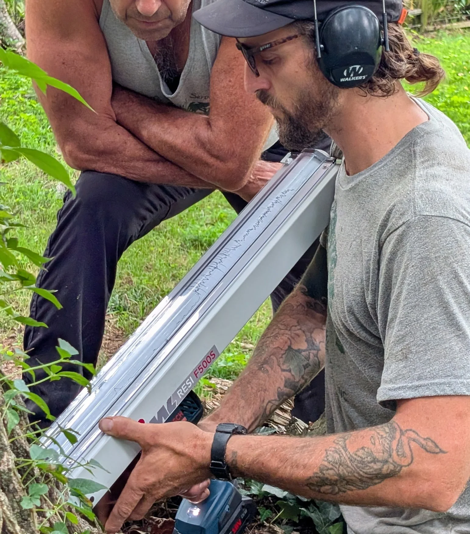 Two men working outdoors, near bushes and greenery. One man has tattoos on his arm and is wearing a gray t-shirt and a watch, while the other man is wearing a gray sleeveless shirt, glasses, and headphones. - Woodwitch Arborists